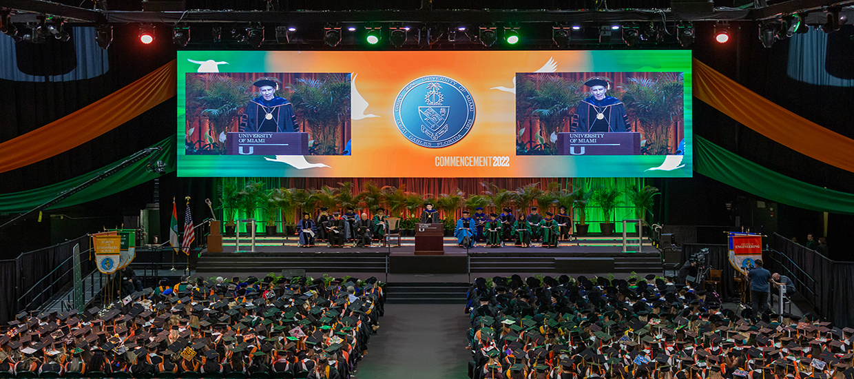 This is an aerial view of the 2022 University of Miami Commencement stage. The university president is speaking at the podium, and the university Dean's are sitting behind him. All of the graduate students are sitting in their chairs facing the stage.